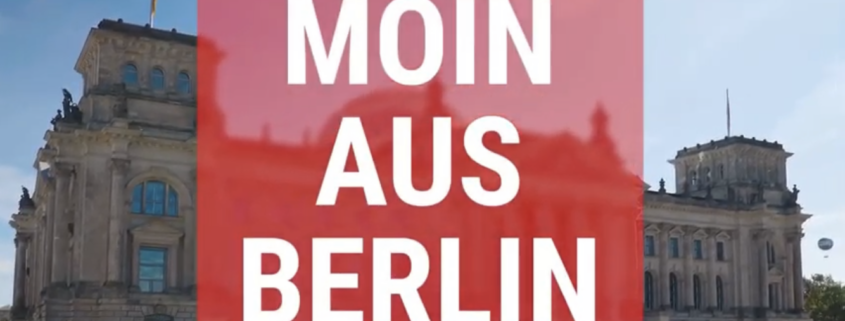 Foto vom Bundestagsgebäude mit weißer Schrift auf rotem Grund "Moin aus Berlin"